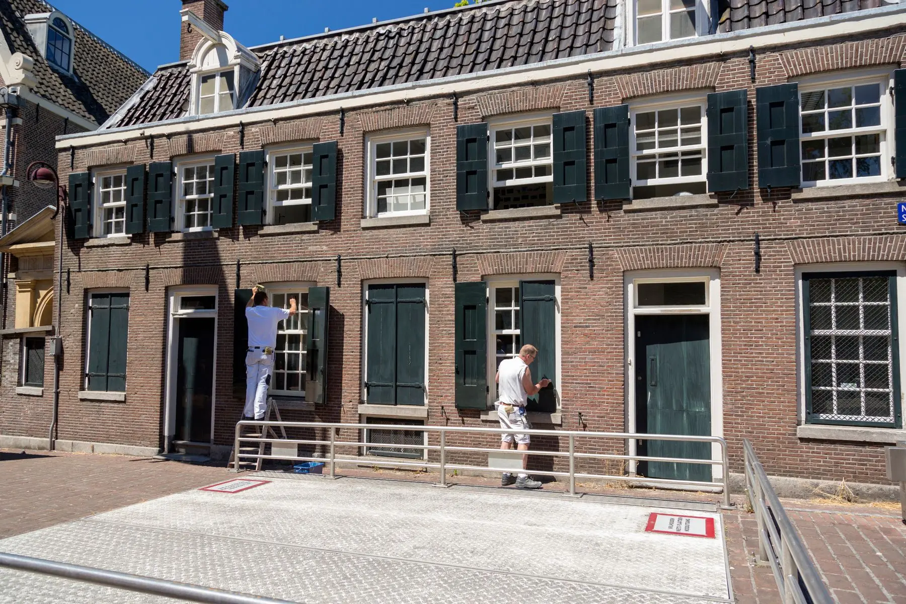 Twee schilders schilderen de kozijnen en luiken van een bakstenen gevel